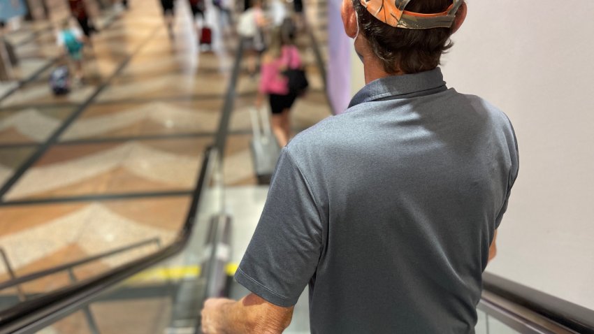 Rear view of senior man wearing cap and mask going down escalator in Denver airport with luggage.  Portrait taken during covid 19  pandemic
